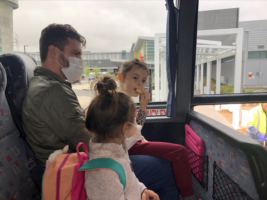 Brandon, Elena, and Eva waiting inside a coach bus. Elena is eating a snack and Eva is wearing a mask.