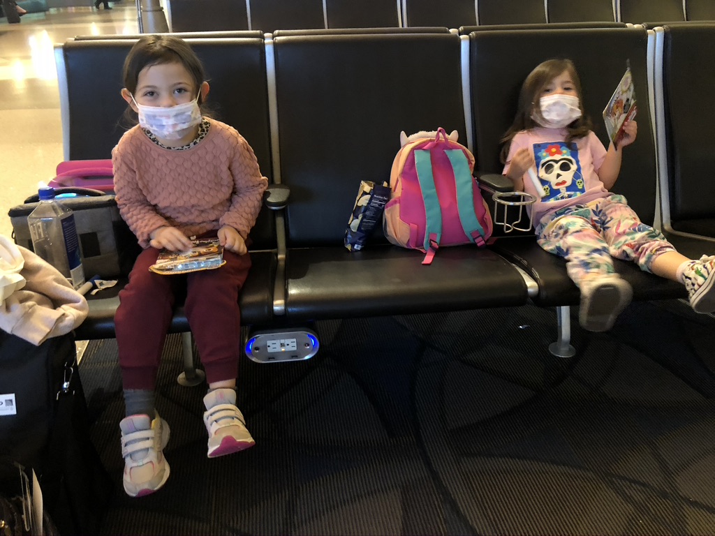 Elena and Eva wearing masks sitting inside an airport waiting to board a flight. They are coloring and eating snacks