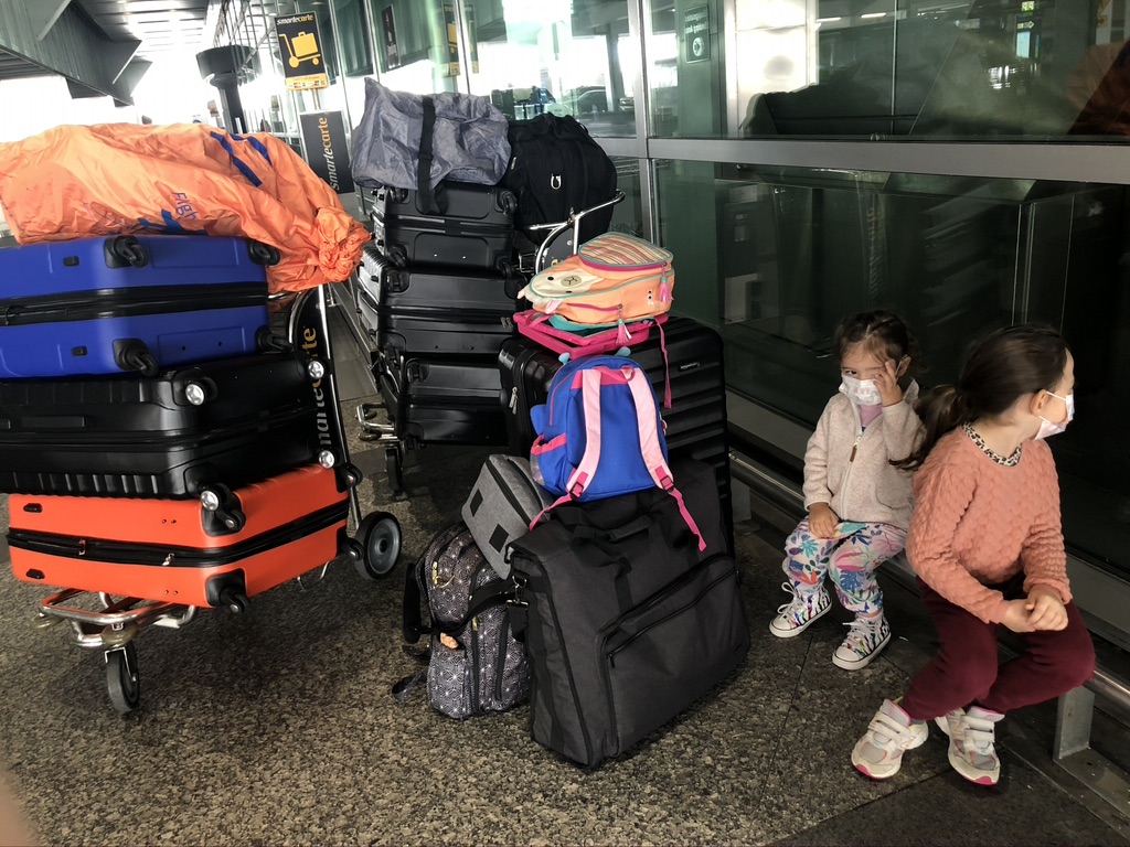 2 small girls sitting in front of an airport wearing masks. Next to them is a mound of luggage.