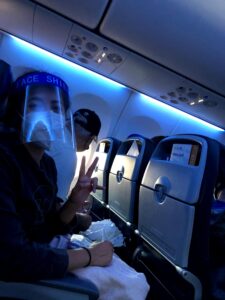 Woman sitting inside of an airplane wearing mask and face shield holding up a peace sign with hand. 