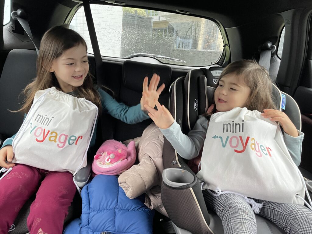 two little girls giving each other a high five in a car before a road trip with their mini voyager travel kit