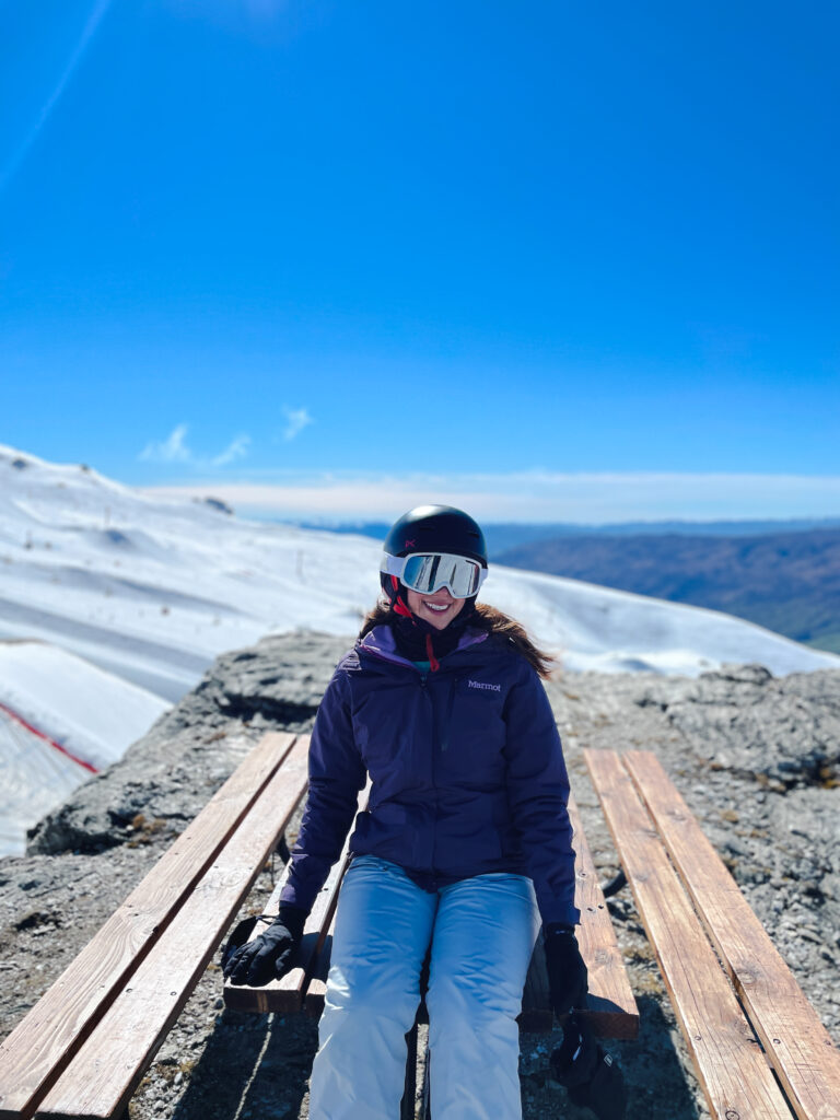 woman sitting on picnic table on Cardrona ski field wearing ski gear with snow covered mountains in the background