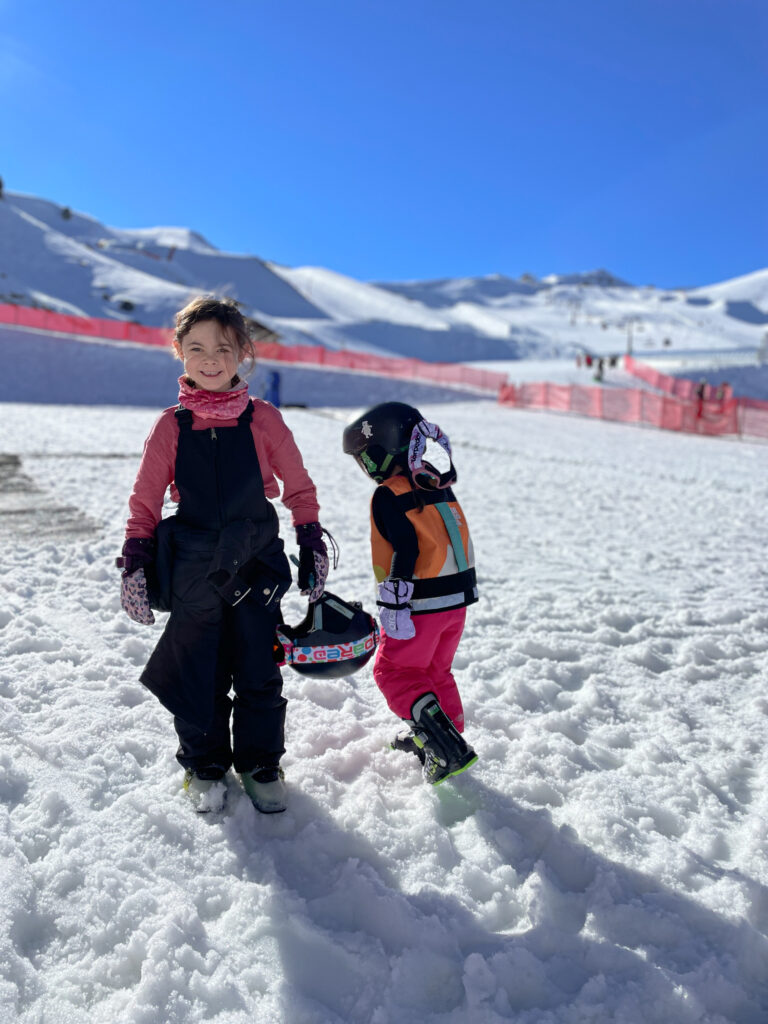 two little girls in full ski gear smiling with snow covered mountain in the background